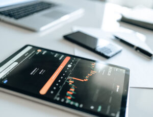 Tablet mit geöffneter Börsen- oder Trading-App auf einem Schreibtisch, im Hintergrund ein Laptop, Smartphone und Stift